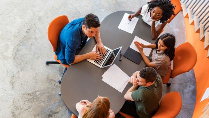 An overhead view of a diverse group of five people sitting around a circular table. They appear to be in a meeting or collaborative workspace, with one person using a laptop and others having papers in front of them.
